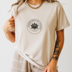Può includere: T-shirt beige con grafica circolare sul davanti con la scritta "Aligned with the Universe" attorno a un disegno di fiore di loto. Il testo "est. within" è sotto il fiore. La maglietta ha maniche corte e girocollo.