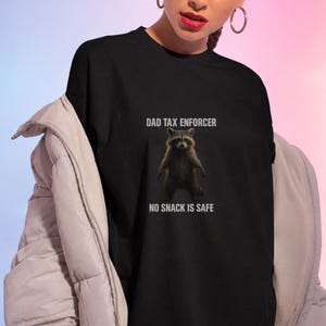 Op de afbeelding: Een zwart t-shirt met een afbeelding van een staande wasbeer. Boven de wasbeer staat de tekst DAD TAX ENFORCER en onder de wasbeer staat de tekst NO SNACK IS SAFE. Het t-shirt wordt gedragen onder een lichtgekleurde, gewatteerde jas met lange mouwen.