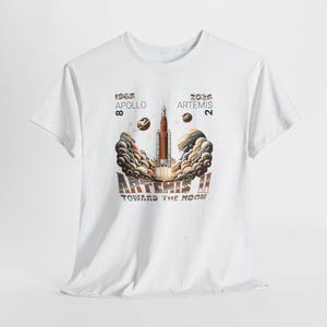 May include: White t-shirt featuring a graphic of a rocket launching with the text "ARTEMIS II TOWARD THE MOON". The design includes the years 1968 and 2026, with the words "APOLLO 8" and "ARTEMIS 2". The rocket is orange and brown.