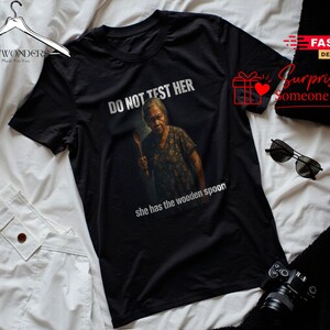 Può includere: Una t-shirt nera a maniche corte con la grafica di una persona anziana che tiene in mano un cucchiaio di legno. Il testo sopra la grafica recita DO NOT TEST HER e il testo sottostante recita she has the wooden spoon in lettere bianche.