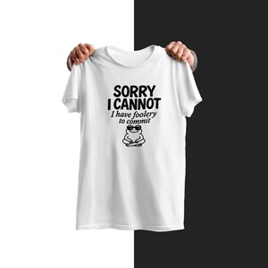 May include: A white short-sleeved t-shirt featuring black text that reads SORRY I CANNOT I have foolery to commit, positioned above a graphic of a small frog wearing sunglasses and sitting in a cross-legged position on the chest area.