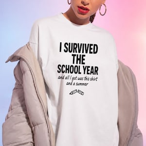 Può includere: T-shirt bianca con la scritta "I SURVIVED THE SCHOOL YEAR" in nero. La maglietta include anche la frase "and all I got was this shirt and a summer" con grafiche di matite. Fa parte di una collezione scolastica.