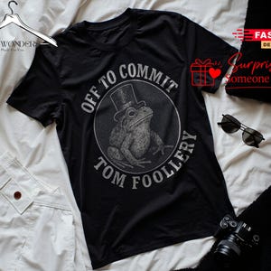 Könnte beinhalten: Ein schwarzes Kurzarm-T-Shirt mit einer kreisförmigen Grafik eines Frosches, der einen Zylinder trägt. Der Text "OFF TO COMMIT TOM FOOLLERY" ist in einer kreisförmigen Anordnung um die Froschillustration in verwaschenem Weiß gedruckt.