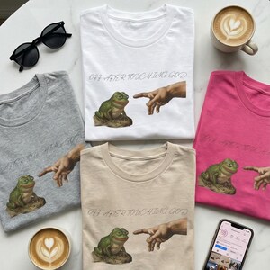 Puede incluir: Camisetas de cuello redondo en blanco, gris, beige y rosa, cada una con un gráfico de una rana y una mano, con el texto "OH AFTER TOUCHING GOD". También se ven unas gafas de sol negras y una taza de café.