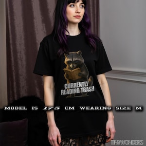 Può includere: Una t-shirt nera a maniche corte con la grafica di un procione che tiene in mano un libro aperto. Sotto il procione, il testo "CURRENTLY READING TRASH" è scritto in lettere maiuscole bianche. Il modello è alto 175 cm.