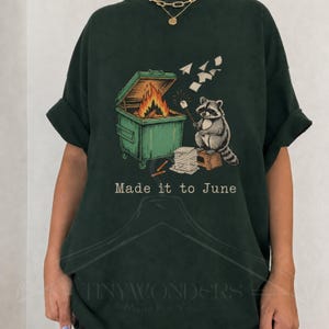 Può includere: T-shirt verde scuro con un procione che arrostisce un marshmallow sopra un fuoco in un cassonetto. La grafica include il testo "Made it to June". La maglietta ha una vestibilità ampia.