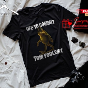 Può includere: Una t-shirt nera a maniche corte con la grafica di una rana verde che indossa un cilindro. Il testo sopra la rana recita OFF TO COMMIT e il testo sotto la rana recita TOM FOOLERY in lettere maiuscole bianche. La maglia ha un girocollo.