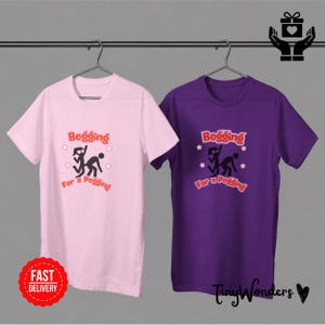 Könnte beinhalten: Zwei T-Shirts, eines rosa und eines lila, hängen an einer Metallstange. Jedes Shirt zeigt den Text "Begging For a Pegging" mit einer Grafik von zwei Strichmännchen. Ein roter "Fast Delivery" Aufkleber befindet sich auf dem rosa Shirt.