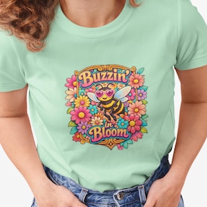 Puede incluir: Camiseta verde claro con un gráfico colorido de una abeja con gafas de sol, rodeada de flores. El texto "Buzzin' in Bloom" se muestra encima y debajo de la ilustración de la abeja. La camiseta es de material suave.