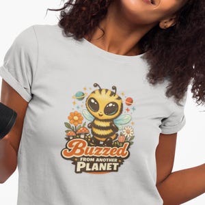 May include: A light gray t-shirt featuring a cartoon bee with large eyes, wearing a black and yellow striped outfit. The shirt has the text "Buzzed From Another Planet" in a retro font, with colorful planets and flowers.