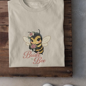 May include: Beige t-shirt with a cartoon bee wearing a veil and holding flowers. The text "Bride to Bee" is printed in pink script below the bee. The shirt is folded on a wooden surface.