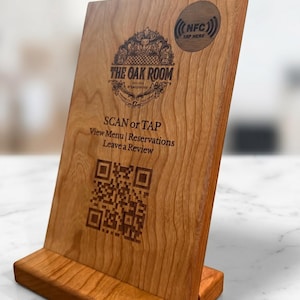May include: A light brown wooden sign with a rectangular base, featuring an engraved logo for The Oak Room, a QR code, and an NFC tap area. The text reads: SCAN or TAP, View Menu | Reservations, Leave a Review, and NFC TAP HERE.