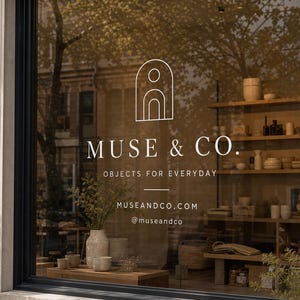 May include: A storefront window displays the white logo and text MUSE & CO. OBJECTS FOR EVERYDAY, MUSEANDCO.COM, and @museandco. Through the glass, wooden shelves hold various ceramic bowls, vases, and woven baskets in a neutral color palette.