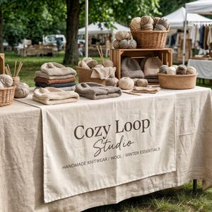 May include: A display of handmade knitwear and wool items at an outdoor market. The table is covered with a beige cloth and features baskets of yarn, knitted hats, and folded sweaters. The sign reads "Cozy Loop Studio."