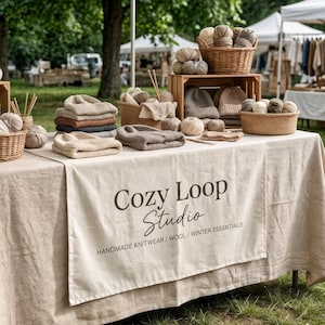 Peut inclure: Un étal de marché présentant des articles en tricot et en laine faits à la main. Des paniers et des caisses en bois exposent de la laine, des bonnets et des tricots pliés dans des tons neutres. Une bannière indique "Cozy Loop Studio".