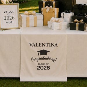 May include: A graduation celebration scene with a table set with gifts, balloons, and floral arrangements. A banner reads "CONGRATS GRAD." The table is decorated with a personalized runner that says "VALENTINA Congratulations! CLASS OF 2026."