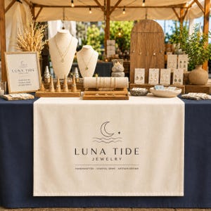 May include: A jewelry display table featuring a cream-colored runner with the Luna Tide Jewelry logo, two necklace busts, wooden earring stands, and ring cones. The display includes various gold necklaces, earrings, and rings on a dark blue tablecloth.