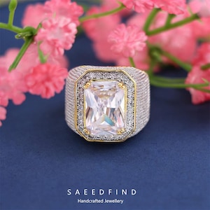 May include: A large, rectangular-cut gemstone ring with a silver band and gold accents. The ring features a prominent, clear central stone surrounded by smaller, sparkling stones. The background includes pink flowers and the text "SAEEDFIND Handcrafted Jewellery."