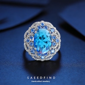 May include: A close-up of a silver ring with a large, oval, light blue gemstone at the center, surrounded by smaller blue stones and intricate detailing. The ring is set against a dark blue background. The text "SAEEDFIND Handcrafted Jewellery" is at the bottom.