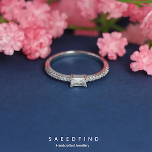May include: A silver ring with a rectangular, clear gemstone at the center, surrounded by smaller, clear stones. The ring sits on a dark blue surface, with pink flowers in the background. The text "SAEEDFIND Handcrafted Jewellery" is at the bottom.