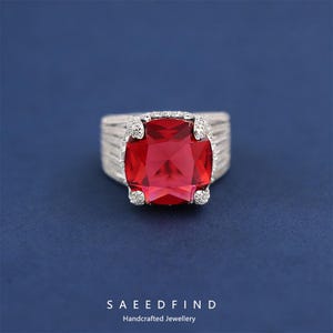 May include: A silver ring featuring a large, square, red gemstone. The ring has a textured band and is embellished with smaller, clear stones around the main gem. The background is a solid, deep blue. The brand name is visible.