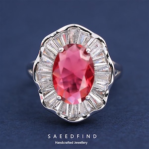 May include: A silver ring featuring a large, oval-cut ruby gemstone surrounded by a halo of baguette-cut white gemstones. The ring has a detailed, ornate design and is set against a dark blue background. The text "SAEEDFIND Handcrafted Jewellery" is at the bottom.