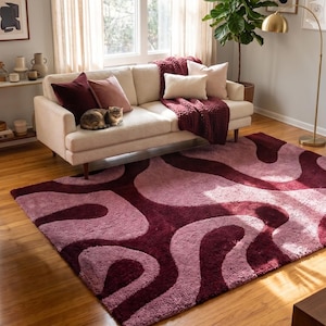 Peut inclure: Salon avec un canapé crème orné de coussins bordeaux et blush, et une couverture assortie. Un tapis à motifs dans les tons bordeaux et rose recouvre le sol. Un chat est assis sur le canapé.