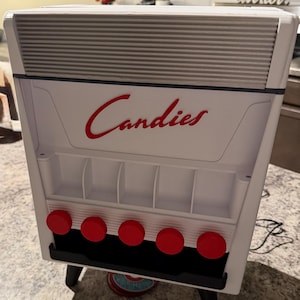 May include: A white candy dispenser with the word "Candies" in red script. The dispenser has a gray top, five red knobs, and a black base. It is supported by black legs.