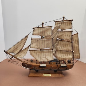 May include: A wooden model of a sailing ship with tan sails and a brown hull. The sails have vertical brown stripes. The ship is mounted on a wooden base with the name "HURRICANE" on a small plaque.