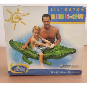 Puede incluir: Caja para un juguete inflable de piscina Lil' Gator Ride-On. El cocodrilo inflable es verde y tiene una cara sonriente. La caja muestra a un hombre y un niño montando el juguete. El juguete mide 168 cm por 86 cm.