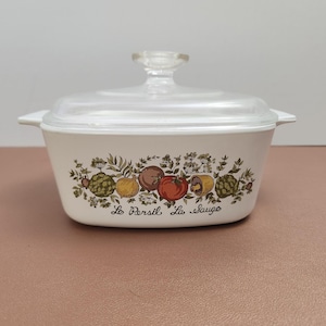 Pode incluir: Travessa branca com tampa de vidro transparente. A travessa apresenta uma faixa decorativa de vegetais coloridos e o texto "Le Persil, La Sauge". A tampa tem uma pega de botão transparente.