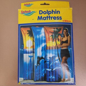 Może przedstawiać: Opakowanie materaca do pływania Dolphin Mattress. Opakowanie zawiera trzy nadmuchiwane materace z motywem delfina i tropikalnego zachodu słońca. Wymiary to około 183 cm x 69 cm.