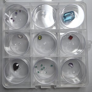 May include: A clear plastic organizer filled with various gemstones in individual compartments. The gemstones are in different shapes, sizes, and colors, including blue, green, red, and purple. The organizer is on a light gray surface.
