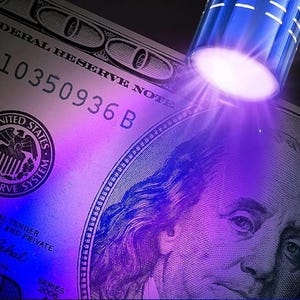 May include: A close-up of a United States $100 bill illuminated by a blue light. The bill displays the text "FEDERAL RESERVE NOTE" and "10350936 B". The image highlights the security features of the currency, with a focus on the portrait of Benjamin Franklin.