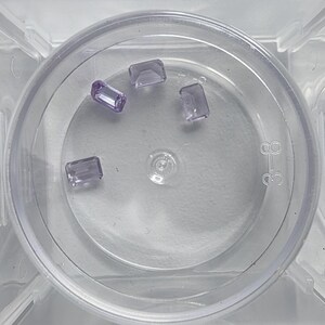 May include: Four rectangular, light purple gemstones are displayed inside a clear, circular container. The gemstones have a polished appearance and are evenly spaced within the container. The container has a ridged edge and a central indentation.