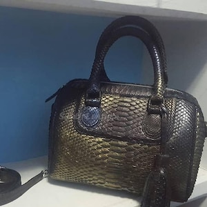 Könnte beinhalten: Eine dunkle, strukturierte Handtasche mit einem oberen Griff und einem abnehmbaren Schultergurt. Die Tasche hat ein Schlangenmuster in Gold- und Schwarztönen. Eine passende Quaste hängt an der Seite. Die Tasche steht auf einem weißen Regal.