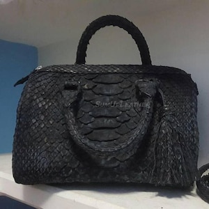 Op de afbeelding: Zwarte handtas met een getextureerd, geschubd patroon, met een handvat aan de bovenkant en een decoratieve kwast. De tas heeft een rechthoekige vorm met een ritssluiting. De tekst "SUMUPLEATHER" is zichtbaar.