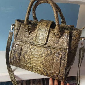 May include: A snakeskin-effect handbag with a top handle and a detachable shoulder strap. The bag has a flap closure and a front pocket. The colour is a mix of olive green, brown, and tan.