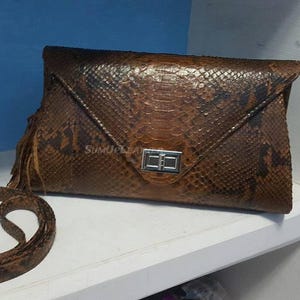 Könnte beinhalten: Braune Schlangenleder-Clutch mit passendem Schulterriemen. Die Clutch hat einen silbernen Verschluss und Fransen. Die rechteckige Tasche hat eine dreieckige Klappe. Der Riemen ist neben der Tasche aufgerollt.