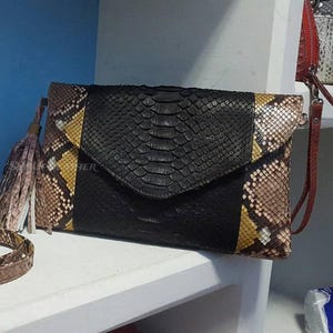May include: A black clutch bag with a flap closure, featuring a snakeskin pattern. The bag has a brown and yellow snakeskin accent on the side and a matching strap. A tassel hangs from the side. The strap is approximately 102 cm long.