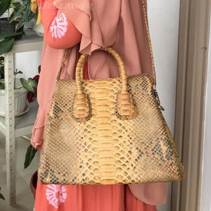 May include: A tan and brown snakeskin handbag with a top handle and a shoulder strap. The bag has a structured shape and a textured surface. The bag is held by a person wearing a pink dress.