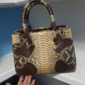 May include: A snakeskin-patterned handbag with a structured design. The bag features a light beige and brown pattern, with a top handle and a rectangular shape. The bag is made of a snakeskin material.
