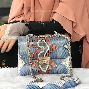 Könnte beinhalten: Eine Handtasche mit blauem, rotem und weißem Schlangenmuster mit goldener Kette und Verschluss. Die Tasche hat eine rechteckige Form und einen passenden gemusterten Riemen. Die Tasche liegt auf einer weißen Oberfläche.