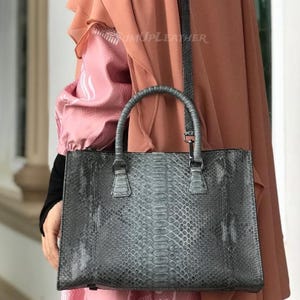 Op de afbeelding: Een grijze handtas met een slangenleerpatroon. De tas heeft een handvat aan de bovenkant en een schouderriem. De tekst "SUMUPLEATHER" is zichtbaar op de tas. De tas is rechthoekig en heeft een ritssluiting.