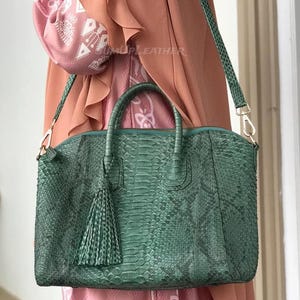 May include: A teal green leather handbag with a snakeskin pattern. The bag features a top handle, a detachable shoulder strap, and a decorative tassel. The bag is held by a person in a pink outfit. The text "MUP LEATHER" is visible on the bag.