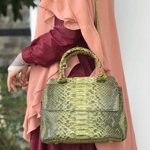 Könnte beinhalten: Eine grüne Handtasche aus Schlangenleder mit einem oberen Griff und einem Schulterriemen. Die Tasche hat einen Klappenverschluss und ein strukturiertes Muster. Der Text "SAMUEL LEATHER" ist auf der Tasche zu sehen. Die Tasche wird von einer Person gehalten, die einen rosa Hijab trägt.