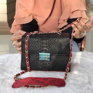 Könnte beinhalten: Schwarze Lederhandtasche mit strukturiertem Muster, mit einem roten und goldenen Kettenriemen. Die Tasche hat einen silbernen Verschluss und ein rotes Innenfutter. Der Markenname "SUMU LEATHER" ist sichtbar.