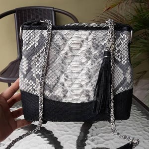 Könnte beinhalten: Handtasche mit Schlangenmuster in Schwarz, Weiß und Grau, mit silberner Kette und schwarzer Quaste. Die Tasche hat eine rechteckige Form mit schwarzem Boden und Klappenverschluss. Der Kettenriemen ist abnehmbar.