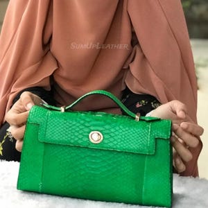 May include: A vibrant green handbag featuring a snakeskin texture. It has a top handle and a flap closure with a gold clasp. The brand name "SUMUPLEATHER" is displayed on the top of the bag.