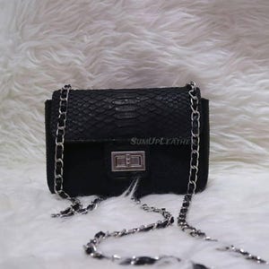 Könnte beinhalten: Eine kleine, schwarze Umhängetasche mit silberner Kette. Die Tasche hat ein strukturiertes, schlangenlederartiges Muster auf der Klappe und dem Körper, mit einem silbernen Verschluss. Der Text "SUMUPLEATHER" ist sichtbar.